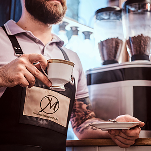 Handsome barista wearing apron drinking coffee during lunch break leaning on a counter in the coffee shop